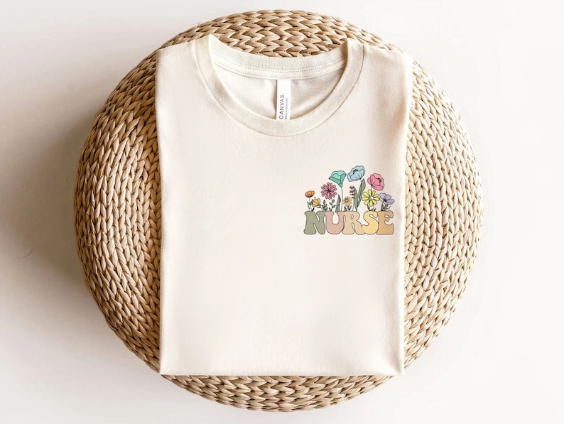 May include: Cream-colored t-shirt with a floral design and the word "NURSE" in a retro font. The flowers are in shades of pink, blue, yellow, and orange. The shirt is made of a soft material and is perfect for everyday wear.