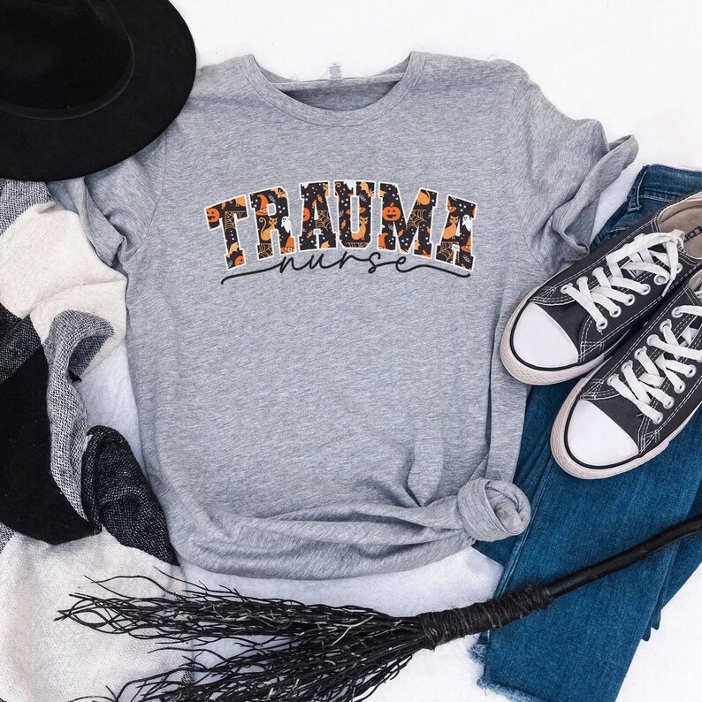 Puede incluir: Una camiseta gris con un gr&aacute;fico de tem&aacute;tica de Halloween que dice "Trauma Nurse" con un patr&oacute;n de calabaza negro y naranja. La camiseta est&aacute; colocada sobre una superficie blanca con una escoba de bruja negra, un sombrero negro y un par de jeans azules con zapatillas negras y blancas.