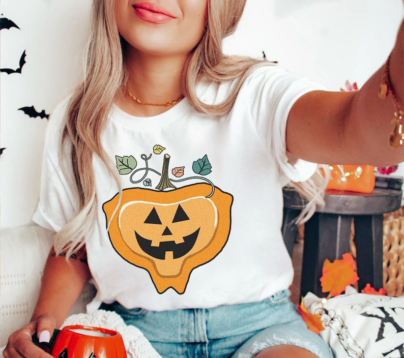 May include: White t-shirt with a graphic of a smiling orange pumpkin with a stem and leaves. The pumpkin is shaped like a heart.