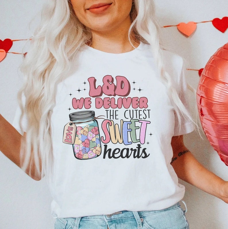 May include: White t-shirt with a pink and white graphic of a mason jar filled with colorful hearts. The text on the shirt reads "L&D We Deliver The Cutest Sweet Hearts".
