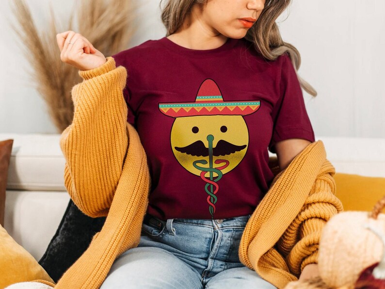 K&ouml;nnte beinhalten: Ein maroonfarbenes T-Shirt mit einem gelben Smiley, der einen bunten Sombrero und einen Schnurrbart tr&auml;gt. Der Smiley hat auch ein gr&uuml;nes und rotes medizinisches Caduceus-Symbol.