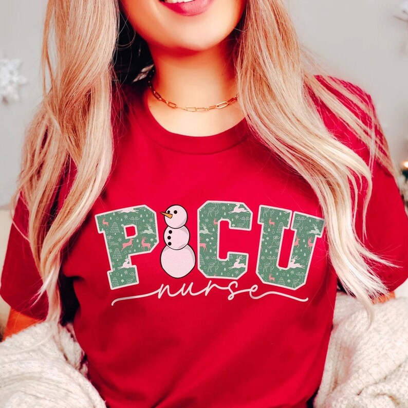 K&ouml;nnte beinhalten: Rotes T-Shirt mit einem gr&uuml;n-wei&szlig;en "PICU nurse" Design, das einen Schneemann mit rosa K&ouml;rper und schwarzem Hut zeigt. Das Design hat ein Weihnachtsmotiv mit kleinen Rentieren und B&auml;umen.
