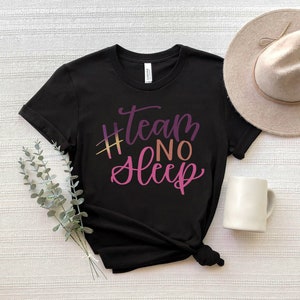 May include: Black t-shirt with the text "#team no sleep" in a gradient of purple, pink, and gold. A beige hat and white mug are next to the shirt. The shirt is folded on a white surface.