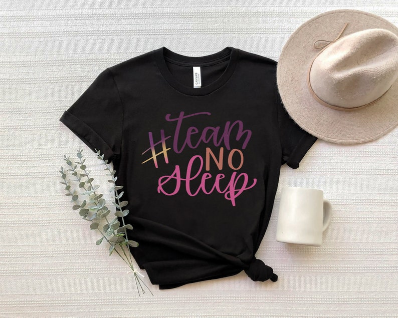 May include: Black t-shirt with the text "#team no sleep" in a gradient of purple, pink, and gold. A beige hat and white mug are next to the shirt. The shirt is folded on a white surface.