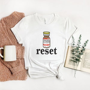 Peut inclure: T-shirt blanc avec un dessin d'un flacon de m&eacute;dicament &eacute;tiquet&eacute; "HALDOL HALOPERIDOL INJECTION, USP" au-dessus du mot "reset" en noir. Le t-shirt est &agrave; c&ocirc;t&eacute; d'un pull marron, d'une tasse blanche et d'un livre ouvert avec de la verdure.
