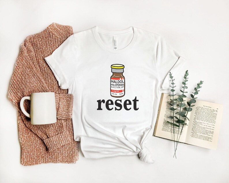 Peut inclure: T-shirt blanc avec un dessin d'un flacon de m&eacute;dicament &eacute;tiquet&eacute; "HALDOL HALOPERIDOL INJECTION, USP" au-dessus du mot "reset" en noir. Le t-shirt est &agrave; c&ocirc;t&eacute; d'un pull marron, d'une tasse blanche et d'un livre ouvert avec de la verdure.