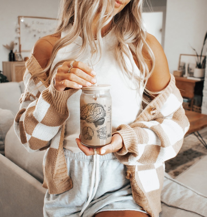 May include: A woman wearing a brown and white checkered cardigan holds a clear glass jar with a straw. The jar has a black and white anatomical illustration of a brain on it.
