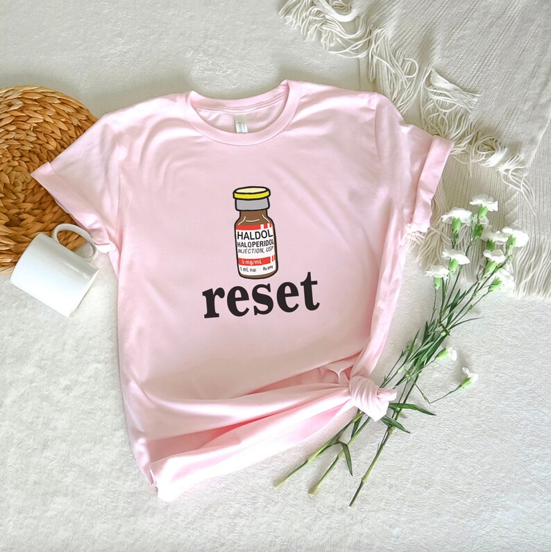 Peut inclure: T-shirt rose clair avec une illustration de flacon de m&eacute;dicament et le texte "HALDOL HALOPERIDOL INJECTION USP". Le mot "reset" est &eacute;crit en noir. Le t-shirt est en mati&egrave;re douce.