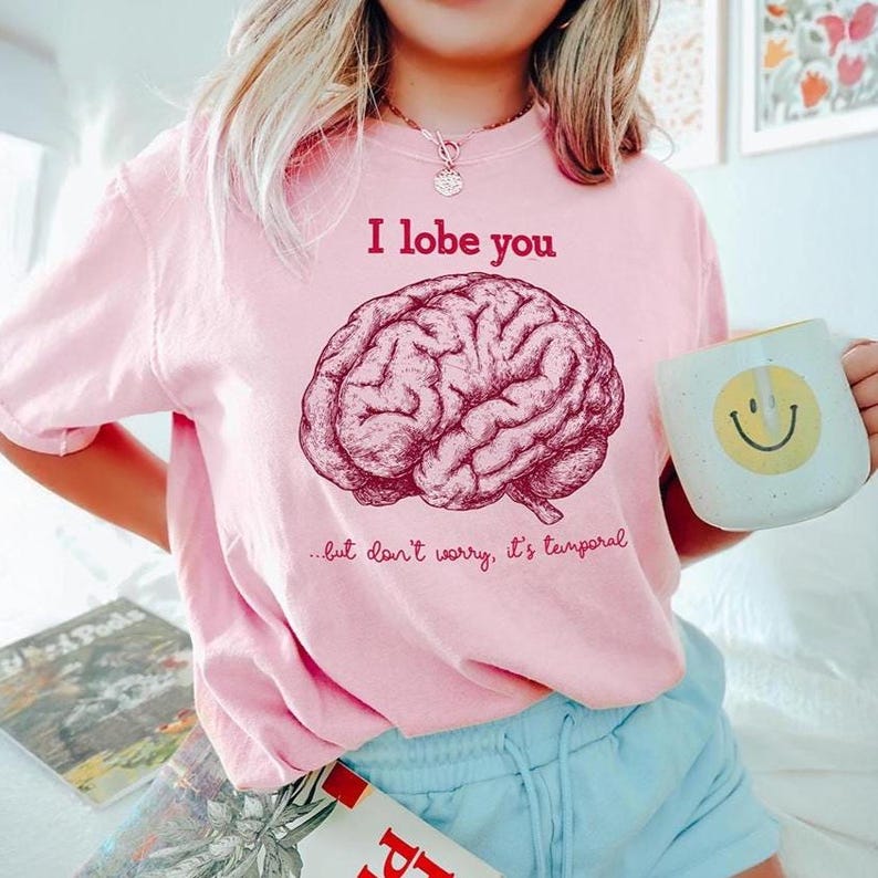 May include: Pale pink t-shirt featuring the words "I lobe you" above a brain illustration. The text "...but don't worry, it's temporal" is below the brain. A white mug with a yellow smiley face is held in the hand.