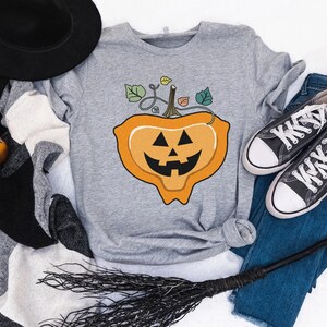May include: A gray t-shirt with a cartoon orange pumpkin with a jack-o'-lantern face. The pumpkin is on a vine with green leaves.