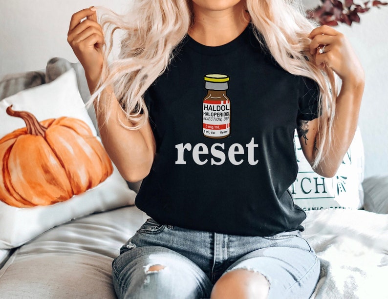 Peut inclure: T-shirt noir avec un motif de flacon de m&eacute;dicament et le mot "reset" en blanc. Le motif du flacon comprend le texte "HALDOL HALOPERIDOL INJECTION USP".