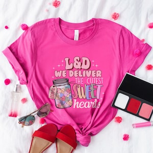 May include: Pink t-shirt with a graphic of a jar filled with colorful hearts and the text "L&D We Deliver The Cutest Sweet Hearts".