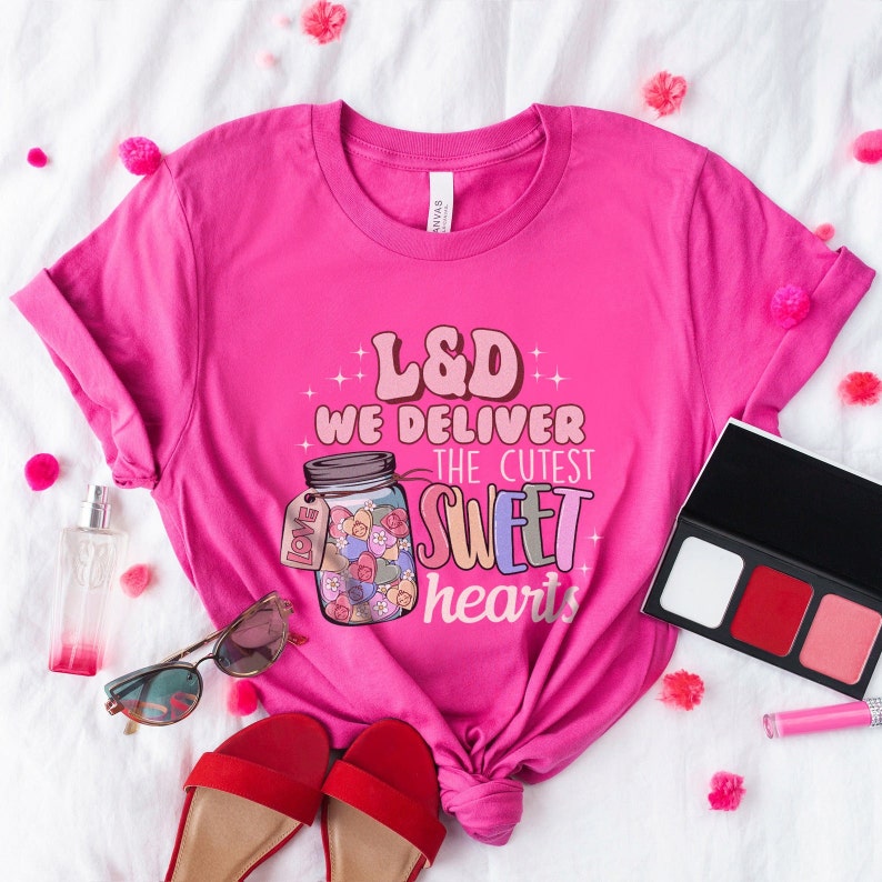 May include: Pink t-shirt with a graphic of a jar filled with colorful hearts and the text "L&D We Deliver The Cutest Sweet Hearts".