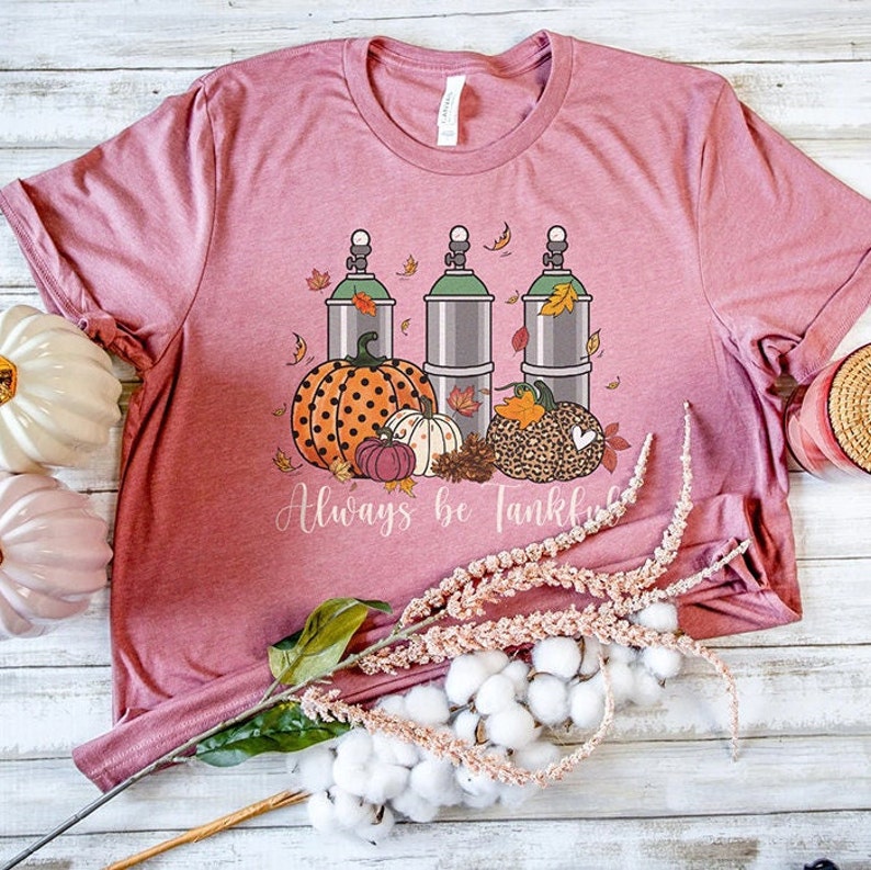 May include: A pink t-shirt with a graphic of three green and silver tanks with fall leaves, pumpkins, and pinecones. The text "Always Be Thankful" is printed below the graphic.