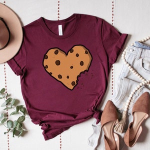 May include: A maroon t-shirt with a brown chocolate chip cookie shaped like a heart with a bite taken out of it.
