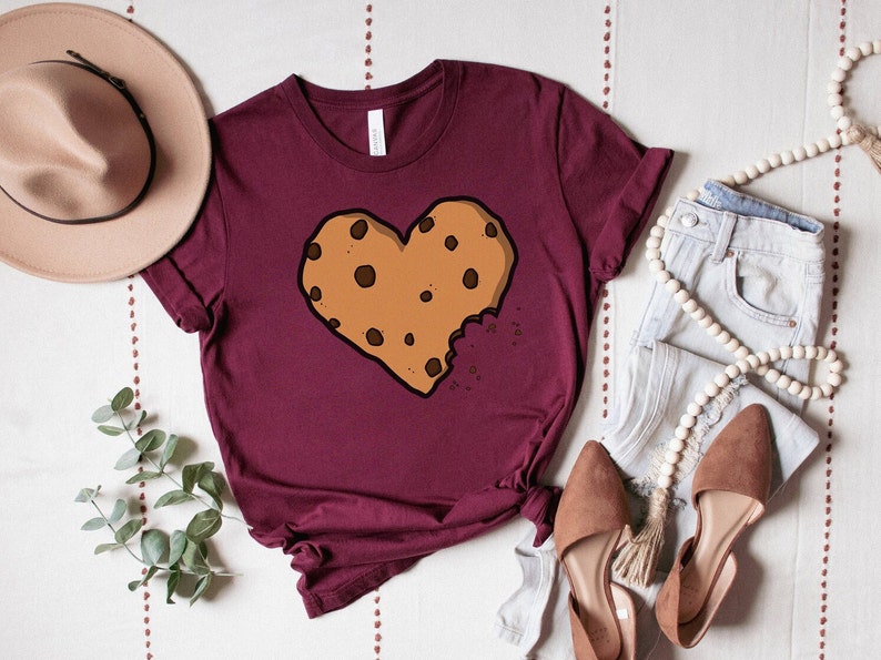 May include: A maroon t-shirt with a brown chocolate chip cookie shaped like a heart with a bite taken out of it.