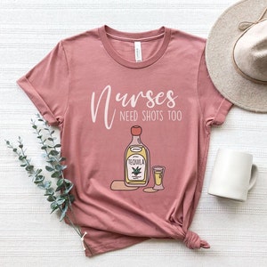 May include: A pink t-shirt with the text "Nurses need shots too" and a cartoon illustration of a bottle of tequila and a shot glass.