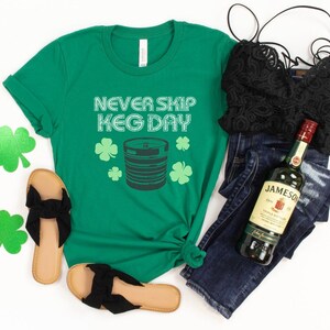 May include: Green t-shirt with the text "Never Skip Keg Day" and a graphic of a beer keg and shamrocks. A bottle of Jameson Irish Whiskey, a pair of black sandals, a black lace bralette, and blue jeans are also pictured.