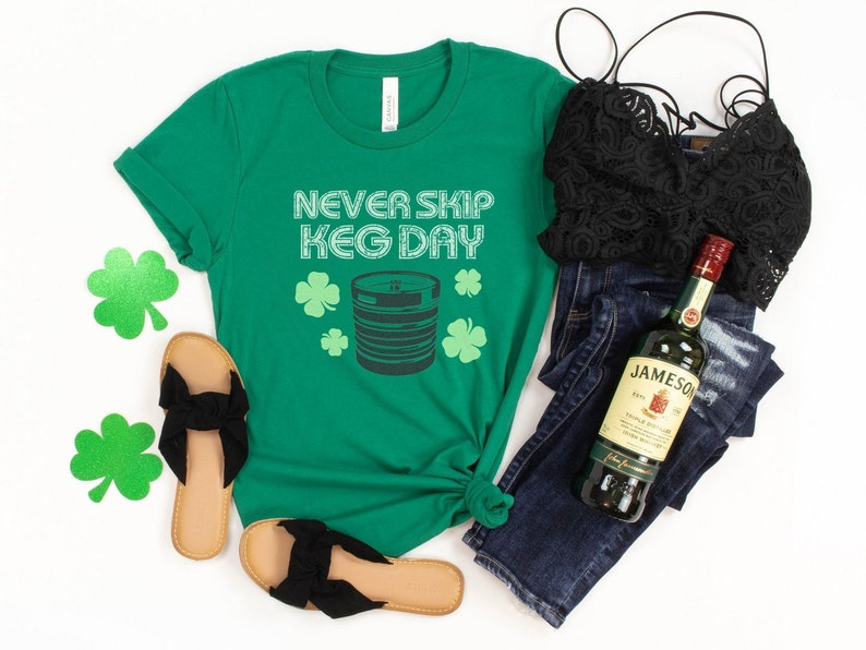 May include: Green t-shirt with the text "Never Skip Keg Day" and a graphic of a beer keg and shamrocks. A bottle of Jameson Irish Whiskey, a pair of black sandals, a black lace bralette, and blue jeans are also pictured.