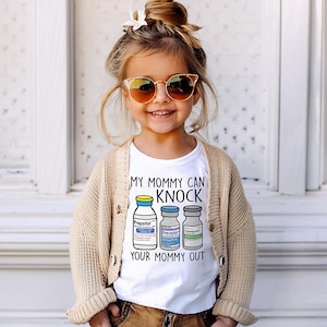May include: A white t-shirt with a graphic of three vials labeled "Propofol", "Precedex", and "Rocuronium". The text on the shirt reads "My Mommy Can Knock Your Mommy Out".