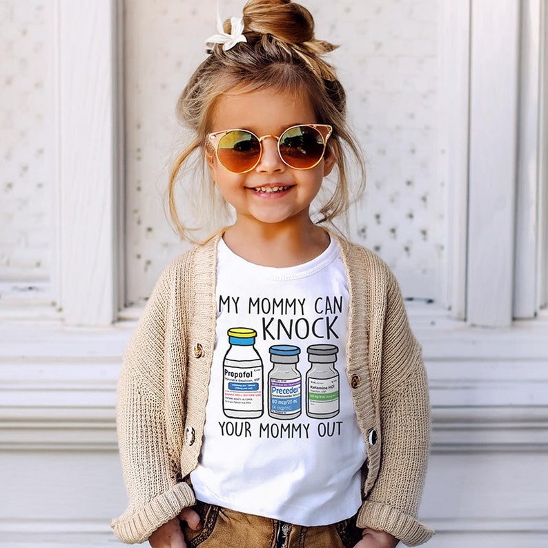 May include: A white t-shirt with a graphic of three vials labeled "Propofol", "Precedex", and "Rocuronium". The text on the shirt reads "My Mommy Can Knock Your Mommy Out".