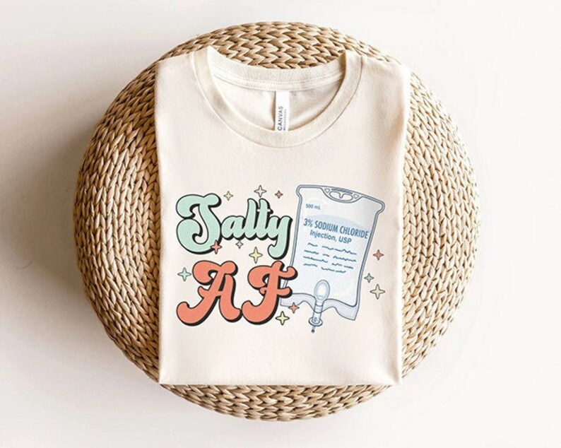 K&ouml;nnte beinhalten: Ein wei&szlig;es T-Shirt mit einem Retro-Grafikdesign, das "Salty AF" mit einer Cartoon-Infusionstasche und Sternen sagt.