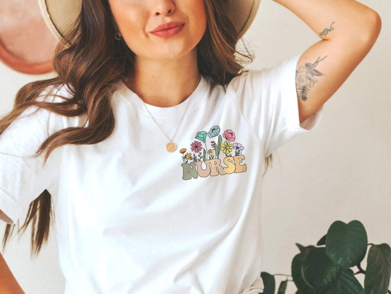 May include: White t-shirt with a colorful floral design and the word "NURSE" in a retro font. The design features various flowers in shades of pink, yellow, blue, and green. The shirt is a casual, everyday wear item.