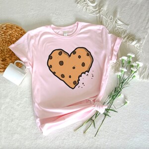 May include: A pink t-shirt with a brown chocolate chip cookie shaped like a heart with a bite taken out of it.