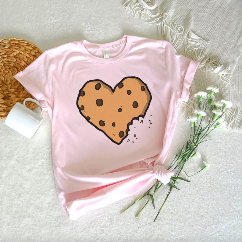 May include: A pink t-shirt with a brown chocolate chip cookie shaped like a heart with a bite taken out of it.