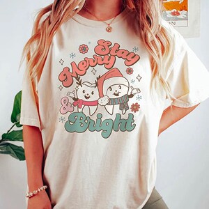May include: A white t-shirt with a Christmas-themed graphic featuring two cartoon teeth wearing Santa hats and the text "Merry & Stay Bright".