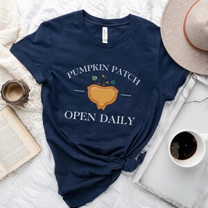 K&ouml;nnte beinhalten: Ein marineblaues T-Shirt mit einer wei&szlig;en Grafik einer K&uuml;rbisplantage mit dem Text "Pumpkin Patch Open Daily".