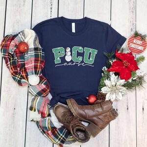 K&ouml;nnte beinhalten: Ein marineblaues T-Shirt mit einem gr&uuml;nen und wei&szlig;en Schneemann-Design. Der Text "PICU nurse" steht unter dem Schneemann. Das Shirt ist von einem karierten Schal, einer roten Weihnachtsstern-Blume und einem kleinen roten Ornament mit der Aufschrift "Frohe Weihnachten" umgeben.