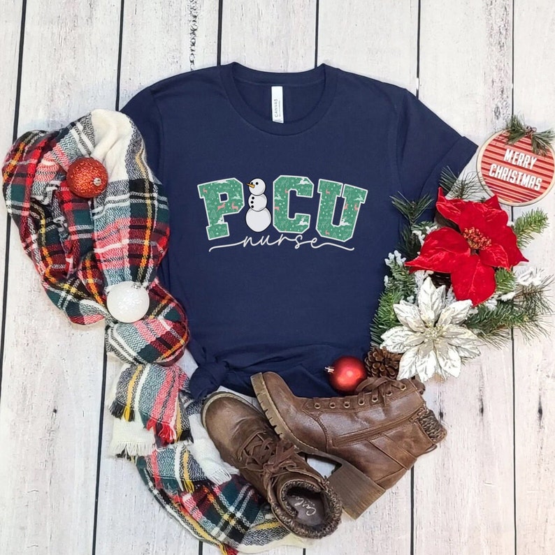 K&ouml;nnte beinhalten: Ein marineblaues T-Shirt mit einem gr&uuml;nen und wei&szlig;en Schneemann-Design. Der Text "PICU nurse" steht unter dem Schneemann. Das Shirt ist von einem karierten Schal, einer roten Weihnachtsstern-Blume und einem kleinen roten Ornament mit der Aufschrift "Frohe Weihnachten" umgeben.