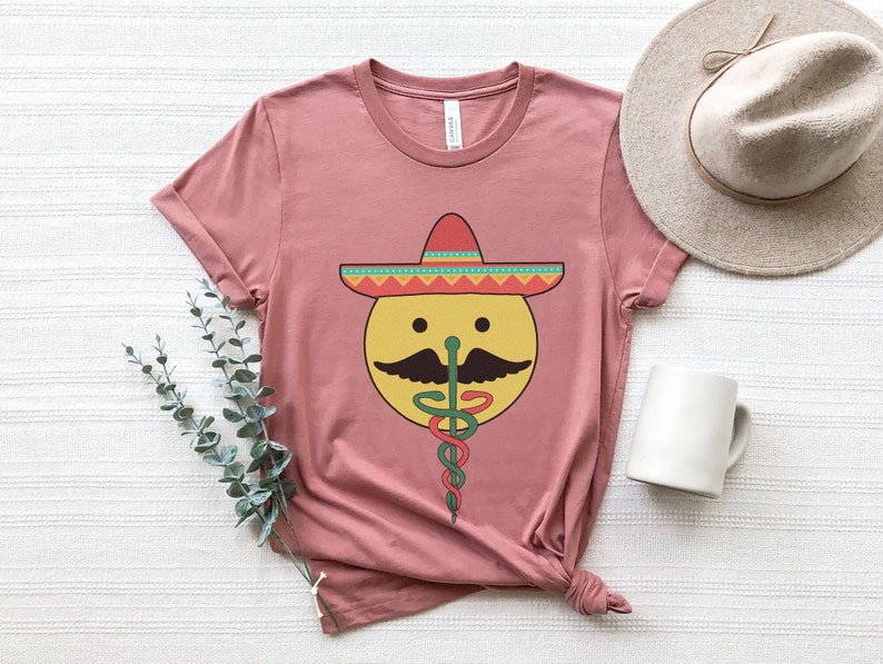 K&ouml;nnte beinhalten: Ein pinkfarbenes T-Shirt mit einem gelben Smiley, der einen roten, gr&uuml;nen und gelben Sombrero tr&auml;gt. Der Smiley hat einen schwarzen Schnurrbart und eine gr&uuml;ne Schlange, die sich um seine Nase windet. Die Schlange ist ein Caduceus, ein Symbol der Medizin.