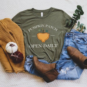 K&ouml;nnte beinhalten: Ein gr&uuml;nes T-Shirt mit dem Text "Pumpkin Patch Open Daily" und einer Grafik einer K&uuml;rbis mit einer Pflanze, die aus ihm herausw&auml;chst. Das Shirt liegt auf einer wei&szlig;en, flauschigen Oberfl&auml;che mit einer blauen Jeans, einem braunen Pullover und braunen Stiefeln.