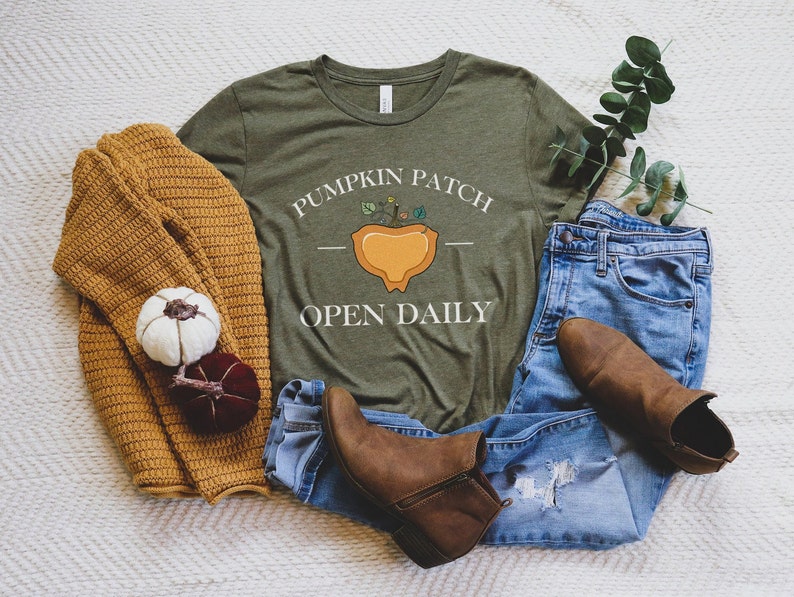 K&ouml;nnte beinhalten: Ein gr&uuml;nes T-Shirt mit dem Text "Pumpkin Patch Open Daily" und einer Grafik einer K&uuml;rbis mit einer Pflanze, die aus ihm herausw&auml;chst. Das Shirt liegt auf einer wei&szlig;en, flauschigen Oberfl&auml;che mit einer blauen Jeans, einem braunen Pullover und braunen Stiefeln.