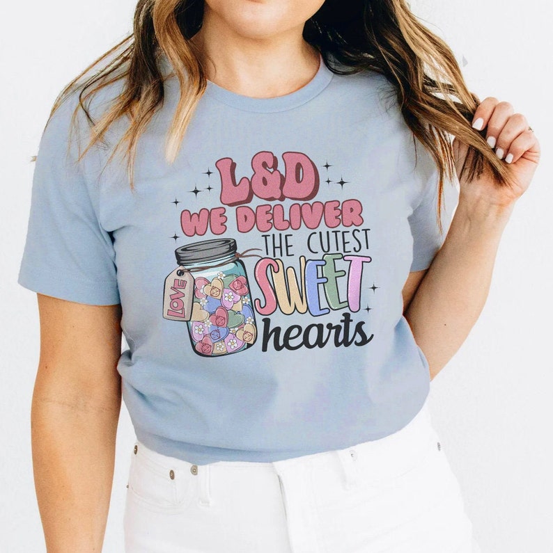 May include: A light blue t-shirt with the text "L&D We Deliver The Cutest Sweet Hearts" and a graphic of a mason jar filled with colorful hearts. The jar has a tag that says "Love".