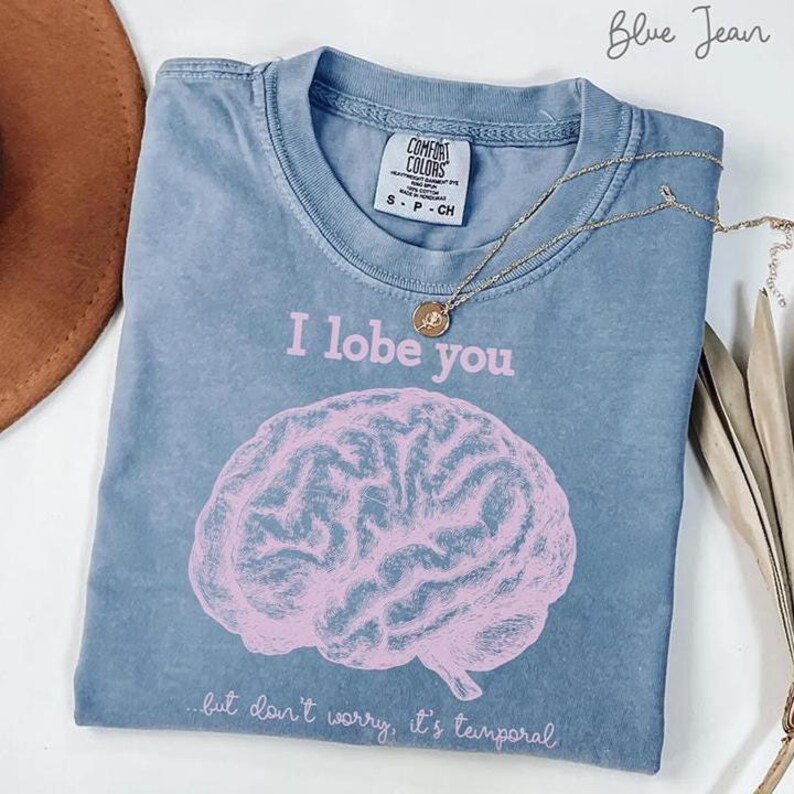 May include: Blue jean coloured t-shirt with the phrase "I lobe you" and a pink brain graphic. The shirt also has the text "...but don't worry, it's temporal." A gold necklace with a pendant rests on the shirt.