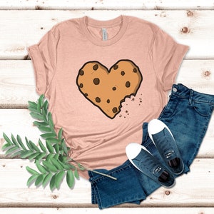 May include: A light pink t-shirt with a brown chocolate chip cookie shaped like a heart with a bite taken out of it. The cookie is on a light pink t-shirt with a white background.