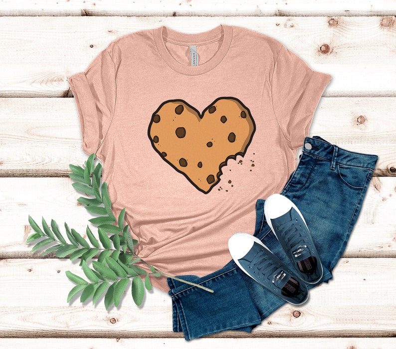 May include: A light pink t-shirt with a brown chocolate chip cookie shaped like a heart with a bite taken out of it. The cookie is on a light pink t-shirt with a white background.