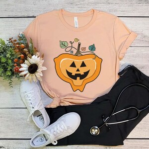 May include: A peach-colored t-shirt with a cartoon jack-o'-lantern design. The jack-o'-lantern is orange with a black face and a green stem with leaves.