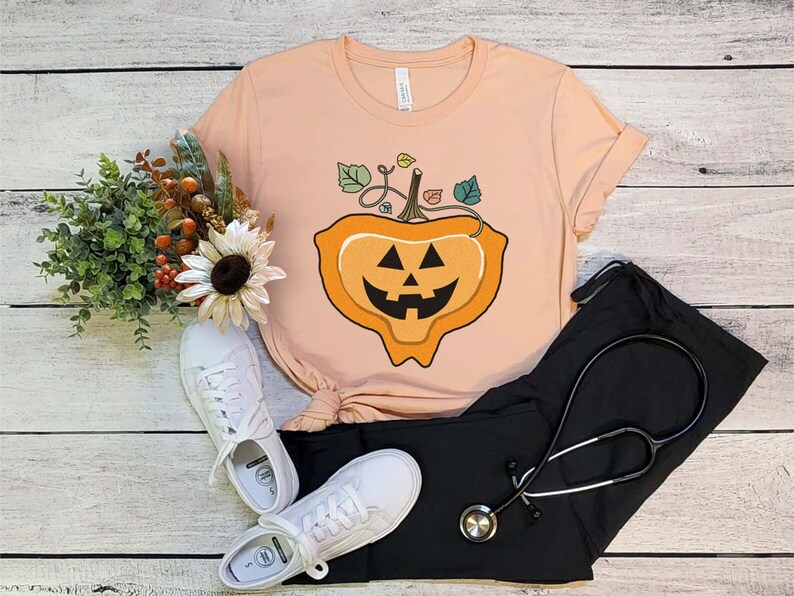 May include: A peach-colored t-shirt with a cartoon jack-o'-lantern design. The jack-o'-lantern is orange with a black face and a green stem with leaves.