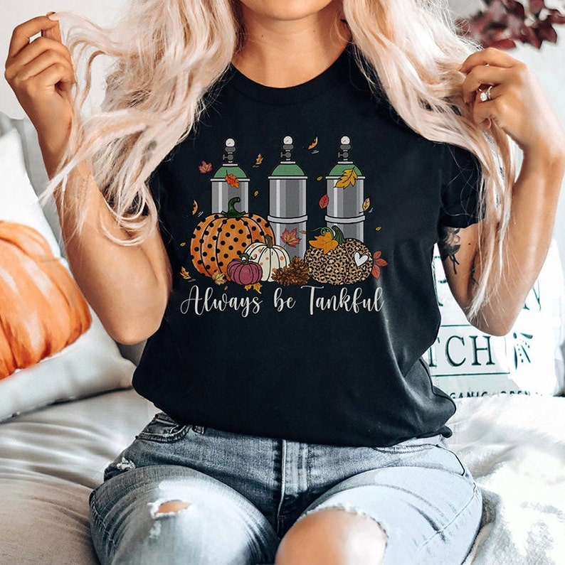 May include: Black t-shirt with a graphic of three green and silver oxygen tanks, pumpkins, and fall leaves. The text "Always be Tankful" is printed below the graphic.