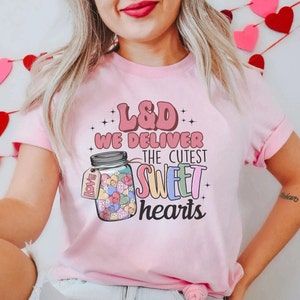 May include: Pink t-shirt with a graphic of a jar filled with colorful hearts and the text "L&D We Deliver The Cutest Sweet Hearts".