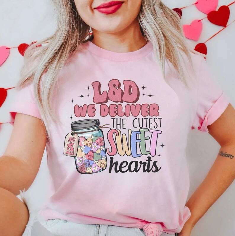 May include: Pink t-shirt with a graphic of a jar filled with colorful hearts and the text "L&D We Deliver The Cutest Sweet Hearts".