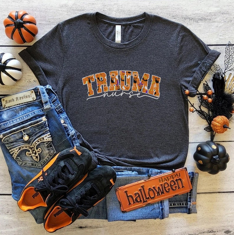 Puede incluir: Una camiseta gris oscuro con un gr&aacute;fico de tem&aacute;tica de Halloween que dice "Trauma Nurse" en letras naranja y negras. El gr&aacute;fico presenta murci&eacute;lagos, fantasmas y calabazas. La camiseta est&aacute; colocada sobre una superficie de madera con jeans, zapatillas y decoraciones de Halloween.