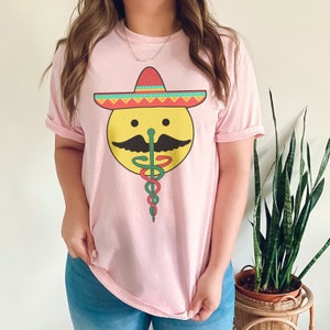 K&ouml;nnte beinhalten: Ein pinkfarbenes T-Shirt mit einem gelben Smiley, der einen roten, gelben und gr&uuml;nen Sombrero tr&auml;gt. Der Smiley hat einen gr&uuml;nen Schlangenbart und einen gr&uuml;nen, roten und gelben Caduceus.