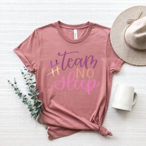 May include: A dusty rose t-shirt with the text "#Team No Sleep" in purple and pink script. A beige hat and a white mug are next to the shirt. A sprig of green leaves is placed near the shirt.