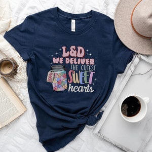 May include: A navy blue t-shirt with a graphic of a mason jar filled with colorful candy hearts. The text "L&D We Deliver The Cutest Sweet Hearts" is printed on the shirt.