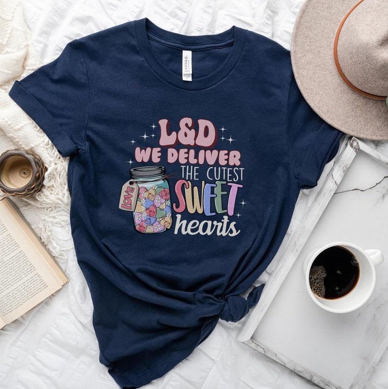 May include: A navy blue t-shirt with a graphic of a mason jar filled with colorful candy hearts. The text "L&D We Deliver The Cutest Sweet Hearts" is printed on the shirt.